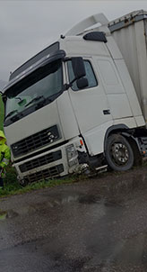 A white Volvo FH truck is overturned on the side of a road. The truck has front-end damage and is partially in a ditch.