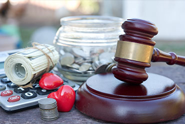 A gavel rests on a sound block, surrounded by a jar of coins, a calculator, a roll of bills, and two red hearts